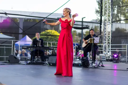 Kobieta w czerwonej sukni śpiewa na scenie, towarzyszą jej muzycy.