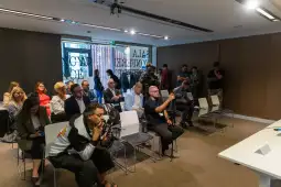 Grupa ludzi siedzących i stojących w sali konferencyjnej, niektórzy robią zdjęcia.