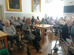 Grupa starszych osób siedzących w sali, część z nich na wózkach inwalidzkich.