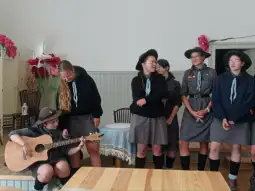 Grupa harcerek w mundurach, jedna z nich gra na gitarze, pozostałe słuchają.