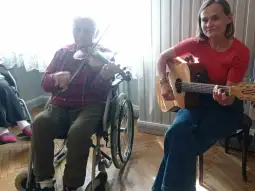 Mężczyzna gra na skrzypcach, a kobieta gra na gitarze. W tle widać innych uczestników.