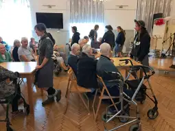 Spotkanie seniorów w świetlicy, rozmowy przy stołach, obecność opiekunów.