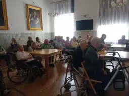 Grupa starszych osób siedzących w sali, część z nich na wózkach inwalidzkich.