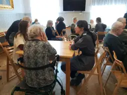 Spotkanie osób starszych z opiekunami w sali z drewnianymi stołami i krzesłami.