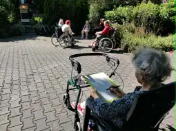 Starsza kobieta rysuje, podczas gdy inne osoby na wózkach inwalidzkich siedzą w parku.