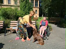 Kobieta w kolorowej sukience głaszcze psa, obok siedzą osoby w wózkach. Słoneczny dzień w parku.