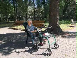 Starsza kobieta siedząca na krześle w parku, obok wózek inwalidzki.
