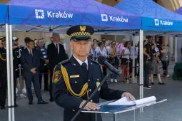Mężczyzna w mundurze stoi przy mównicy, w tle tłum ludzi i namioty z logo Kraków.
