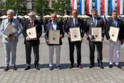 Sześciu mężczyzn trzymających ramki z dyplomami na tle flag i żołnierzy.