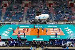 Mecz siatkówki w hali sportowej z drużynami w czerwonych i niebieskich strojach.