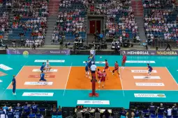 Mecz siatkówki w hali sportowej z zawodnikami w niebieskich i czerwonych strojach.