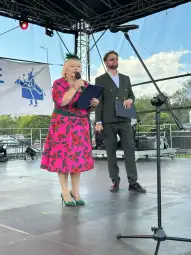 Kobieta w różowej sukience z kwiatowym wzorem przemawia na scenie obok mężczyzny w garniturze.