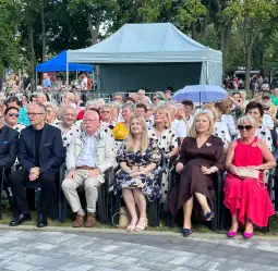 Grupa ludzi siedzących na krzesłach, w tle widoczna publiczność i namiot.