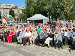 Tłum ludzi siedzący na krzesłach, część z parasolami, w tle zjeżdżalnia dla dzieci.