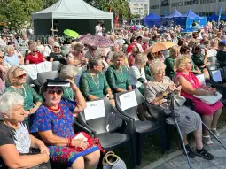 Grupa kobiet w strojach ludowych siedzących na widowni podczas wydarzenia plenerowego.