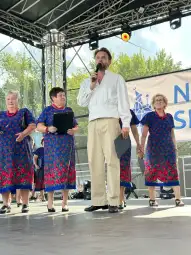 Mężczyzna z mikrofonem na scenie, obok grupy kobiet w kwiecistych sukienkach.