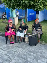 Trzy starsze kobiety w kolorowych kapeluszach siedzą na krzesłach, ubrane w eleganckie stroje.