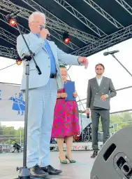 Mężczyzna w jasnym garniturze przemawia do publiczności, obok niego dwie osoby trzymające dokumenty.