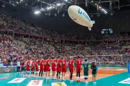 Zawodnicy drużyny siatkarskiej w czerwonych strojach na tle pełnej trybuny.