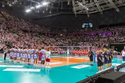 Mecz siatkówki z drużynami w strojach sportowych i kibicami na trybunach.