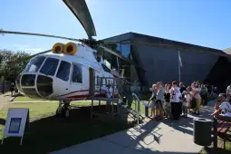 Helikopter wystawiony na zewnątrz, obok grupy ludzi czekających w kolejce.