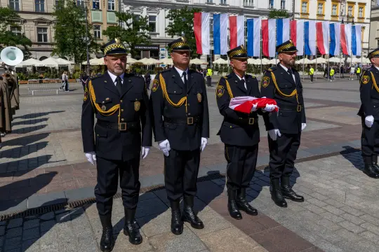 Czterech mężczyzn w mundurach stoi w szeregu, trzymając flagę. W tle dekoracje w narodowych kolorach.