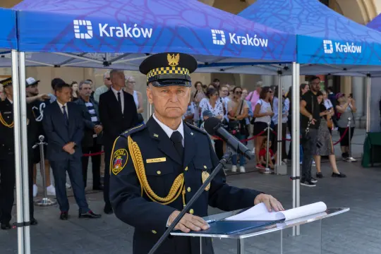 Mężczyzna w mundurze stoi przy mównicy, w tle tłum ludzi i namioty z logo Krakowa.