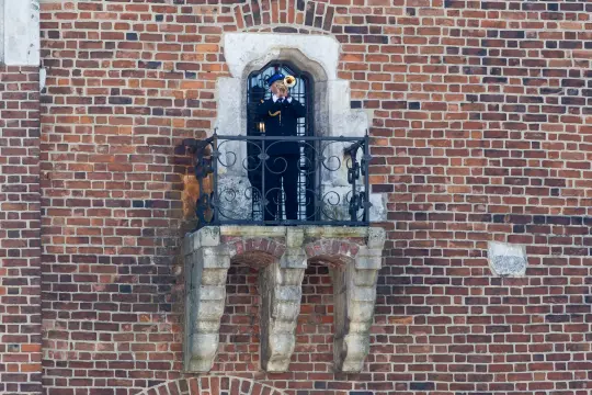 Mężczyzna w mundurze grający na trąbce z balkonu na tle ceglanej ściany.