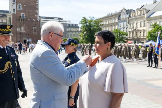 Wręczenie odznaczenia na uroczystości w plenerze, w tle tłum ludzi i budynki.
