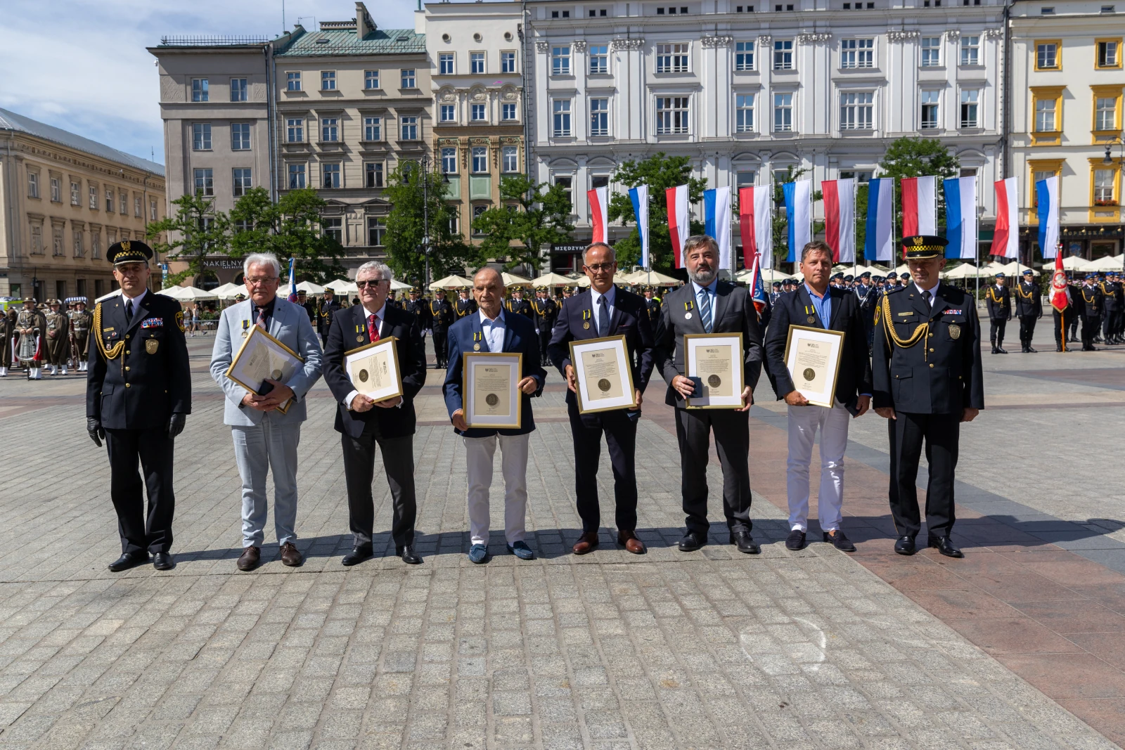 Sześciu mężczyzn w garniturach trzymających dyplomy na tle flag i zgromadzenia ludzi.  Autor: Bogusław Świerzowski
