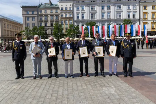 Sześciu mężczyzn w garniturach trzymających dyplomy na tle flag i zgromadzenia ludzi.