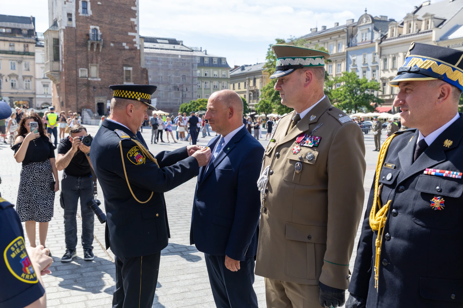 Ceremonia wręczenia odznaczenia w miejskim otoczeniu z uczestnikami i przedstawicielami służb mundurowych.  Autor: Bogusław Świerzowski