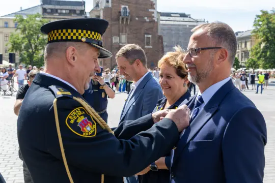 Ceremonia wręczenia odznaczeń w Krakowie, uśmiechnięci uczestnicy w formalnych strojach.