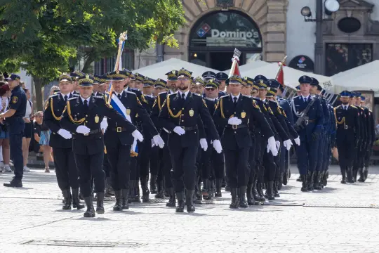 Parada policji w mundurach, z flagami, na ulicy w słoneczny dzień.