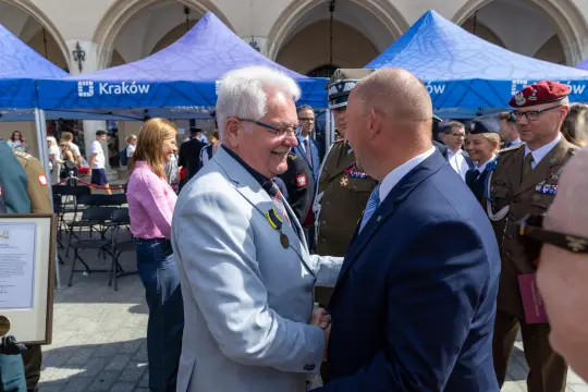Mężczyźni w garniturach uścisną ręce podczas uroczystości na placu. W tle namioty z logo Krakowa.