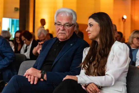 Mężczyzna i kobieta rozmawiają w eleganckim otoczeniu, oboje w formalnych strojach.