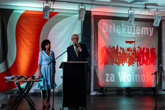 Mężczyzna przemawia przy mównicy, obok kobieta, w tle flagi i hasło Dziękujemy za Wolność.