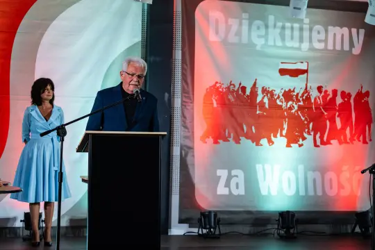 Mężczyzna przemawia przy mównicy, obok kobieta w niebieskiej sukience, w tle hasło o wolności.