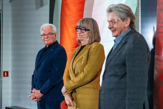 Trzy osoby stojące obok siebie, uśmiechające się, w formalnych ubraniach.