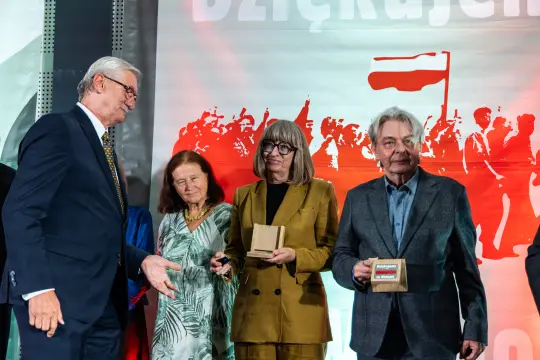 Cztery osoby na scenie podczas uroczystości wręczenia nagród, w tle flagi i tłum.