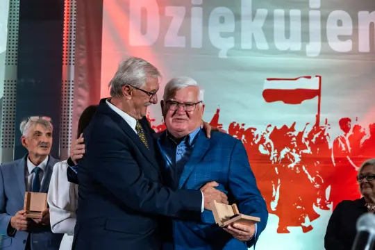 Dwaj mężczyźni w garniturach uścisnęli się, w tle widoczny napis Dziękujemy oraz motyw z flagą Polski.