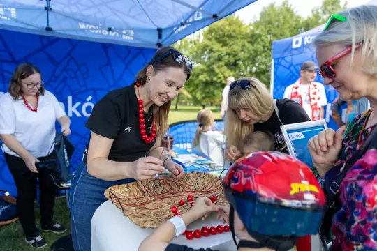 Kobieta w czerwonych koralikach uczy dzieci robienia biżuterii na festynie.