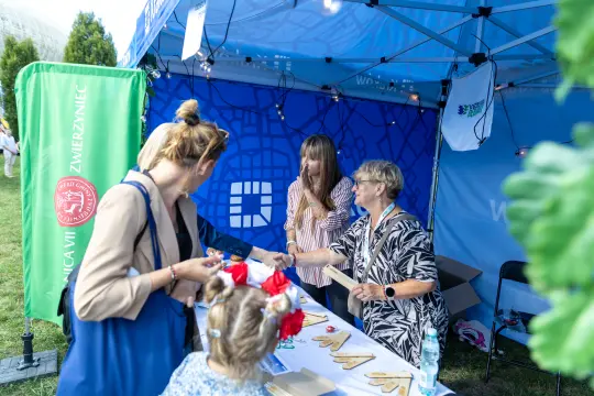 Kobiety przy stoisku, rozmawiają i wymieniają się materiałami promocyjnymi. Dziecko z kokardkami w tle.