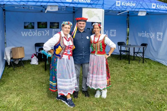 Trzy osoby w tradycyjnych strojach ludowych, uśmiechnięte, w plenerze pod namiotem.