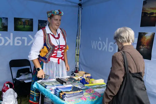 Kobieta w tradycyjnym stroju rozmawia z klientką przy stoisku z materiałami promocyjnymi.