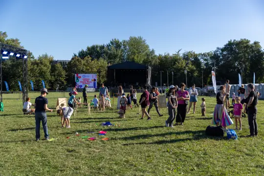 Ludzie bawią się na festynie w parku, w tle scena z występami.