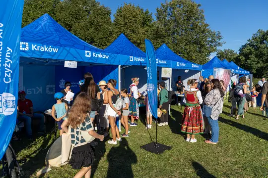 Tłum ludzi przy namiotach promocyjnych w Krakowie, w tle zieleń drzew.