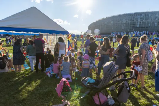 Tłum ludzi na festynie, stoiska z atrakcjami, namiot i stadion w tle.