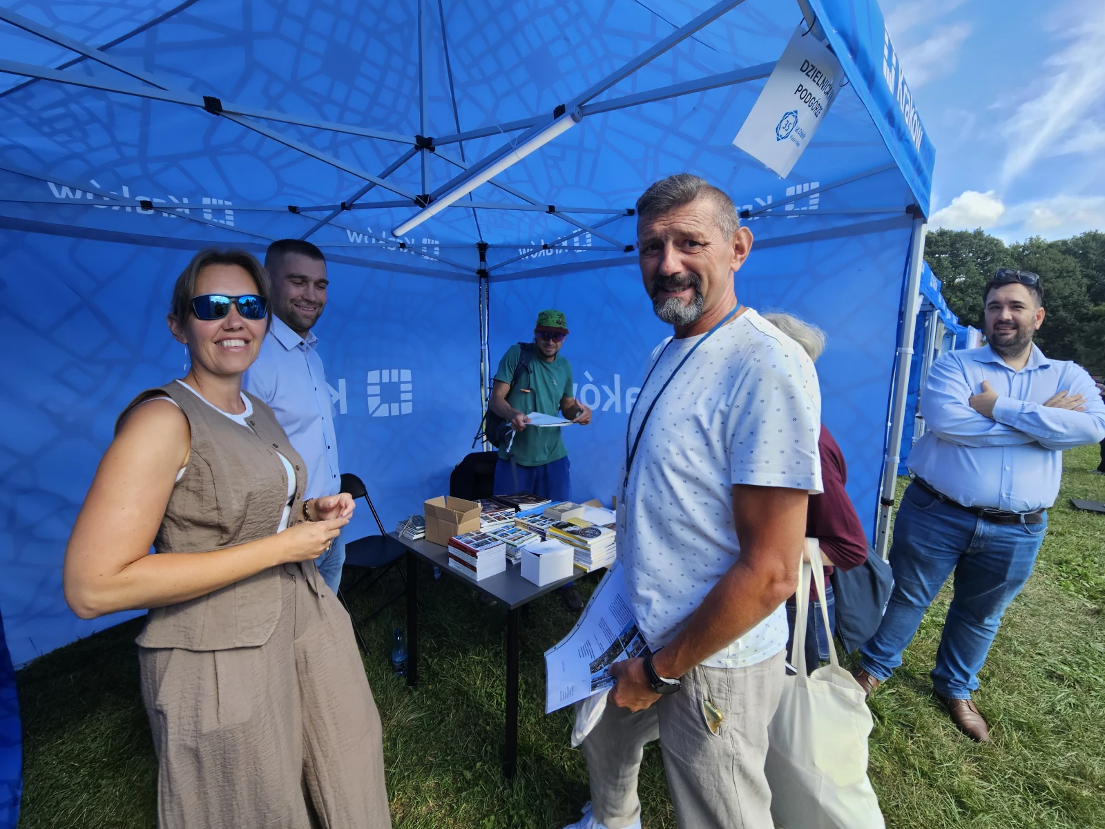 Grupa ludzi przy stoisku z materiałami promocyjnymi na wydarzeniu plenerowym.  Autor: Bogusław Świerzowski