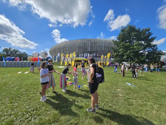 Dzieci bawią się na trawie przed dużym budynkiem, w tle balony i flagi.